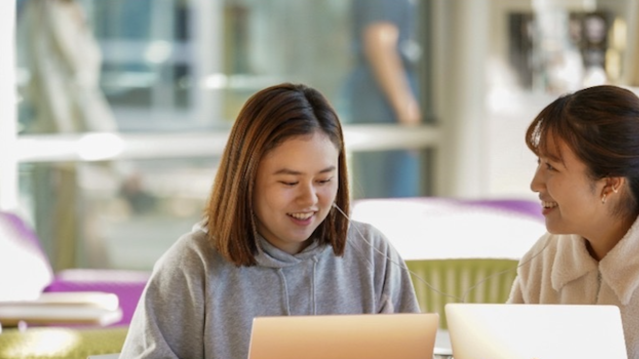 Two young women smile and engage while working on laptops in a bright, modern space.