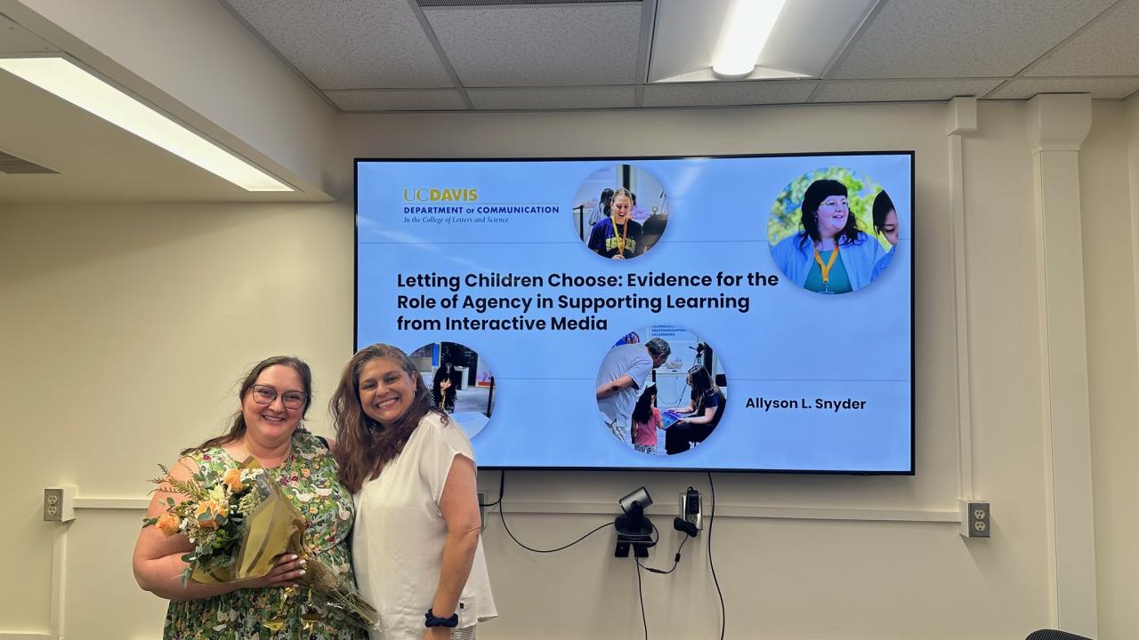 Stephanie Fallas (right) smiling with a student (left) in a conference room in front of a presentation screen.