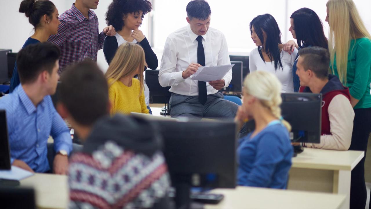 group of older students standing around, listening intently to a faculty member who wearing a white shirt and tie in a computer room. The faculty member's head is down as he's pointing to a piece of paper in his hand,