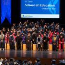 Graduation ceremony on auditorium stage with faculty in academic regalia and graduates, blue banners