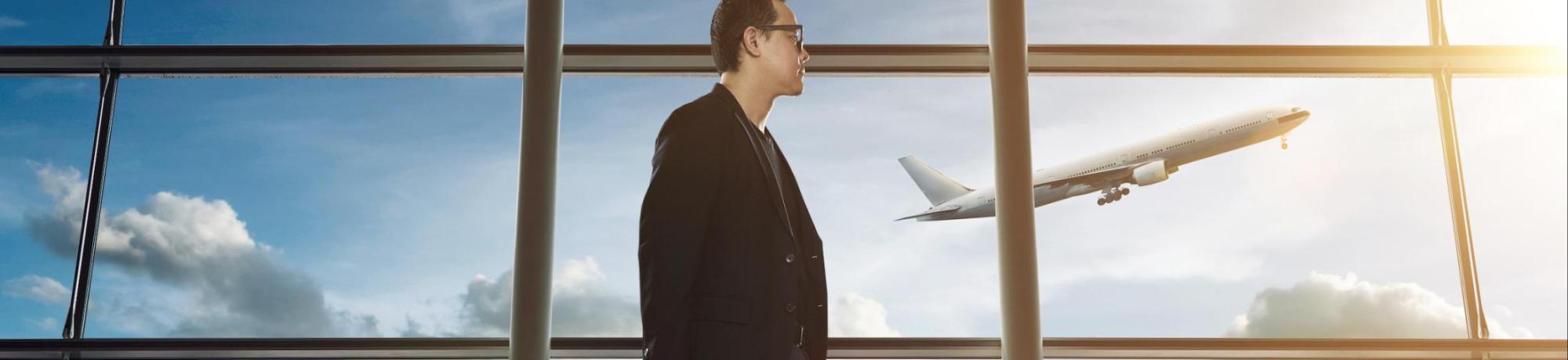 side profile of a man walking through an airport in suit. through large windows behind him a plane is taking off off into rising sun